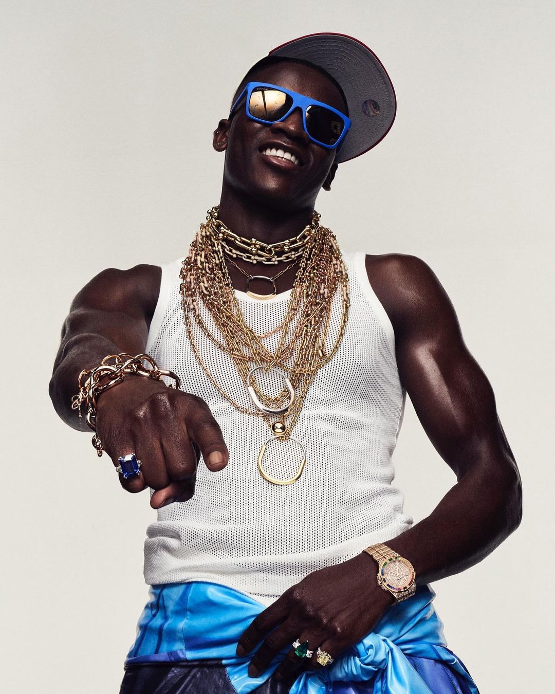 Portrait of a dark-skinned adult man with a backwards baseball cap, sunglasses, and multiple gold chains posing against a white background.