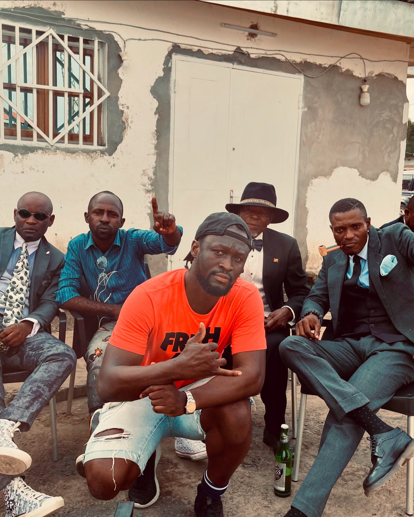 Group of six African men pose outdoors, some sitting on chairs, one squatting, wearing suits, hats, and casual clothes, against a building backdrop.