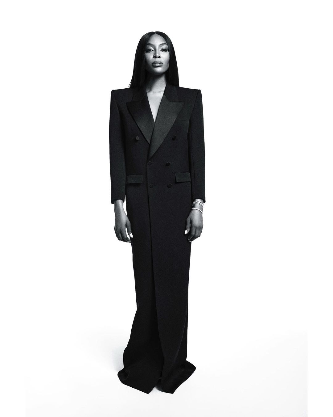 Black and white portrait of a black female model wearing a long black double-breasted jacket dress against a clean white backdrop.