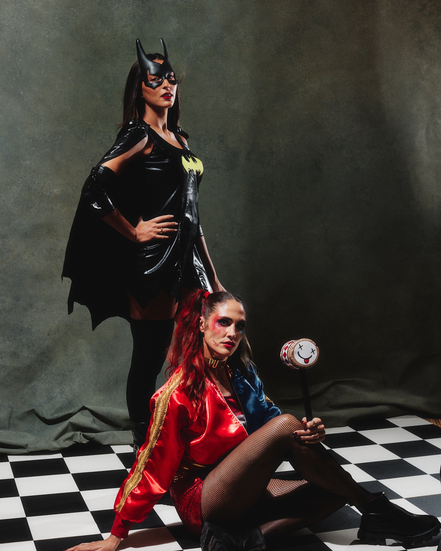 Two women pose in costume, one as Batgirl in a black vinyl dress and mask, the other as Harley Quinn with red and blue hair.