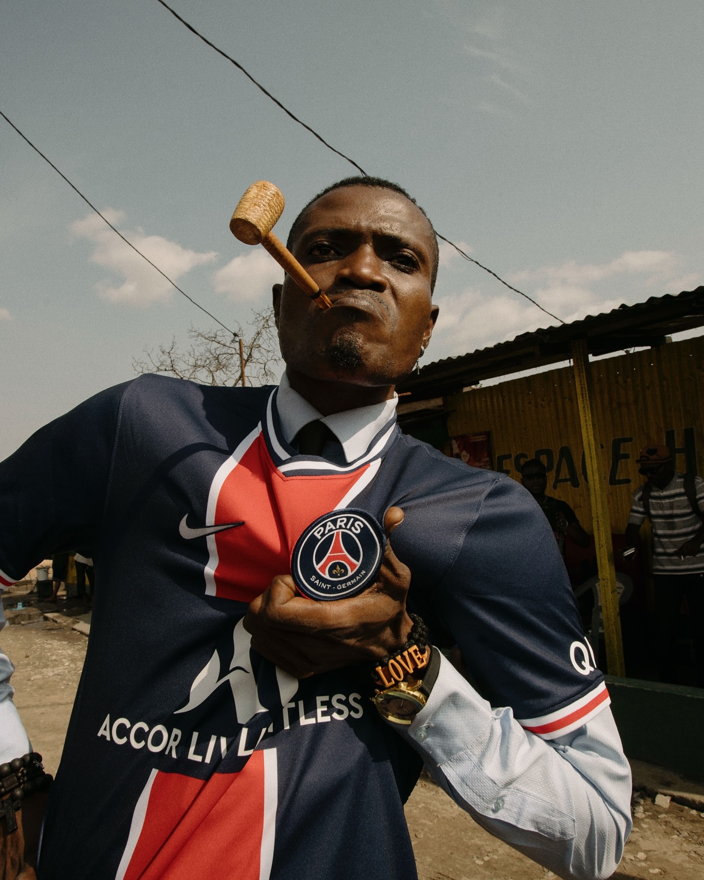 Adult male in sports jersey holds Paris Saint-Germain emblem outdoors, smoking a wooden pipe against a bright sky background.