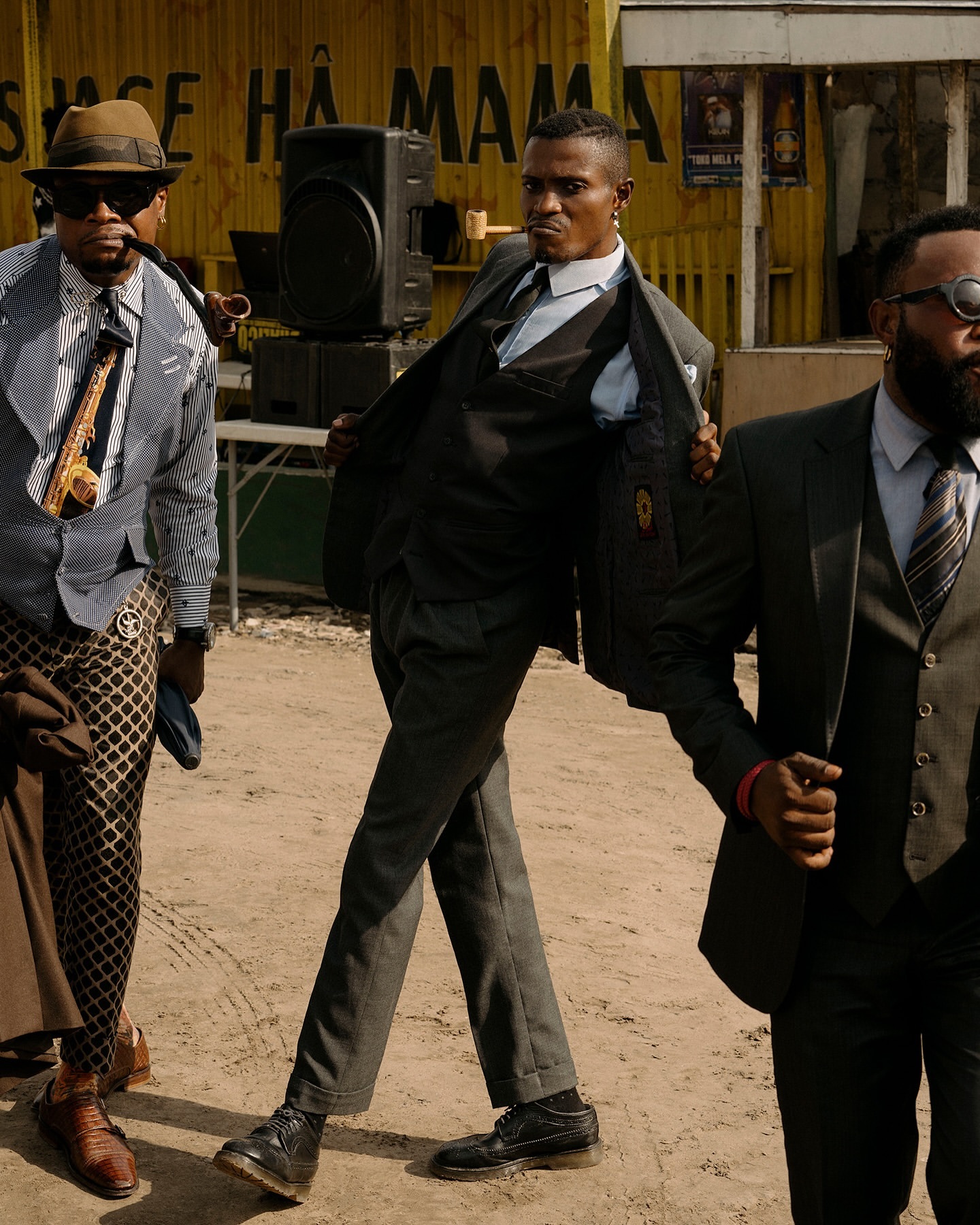 Three adult males in suits pose outdoors, one with a pipe, another with an open jacket, against a backdrop of a yellow structure.