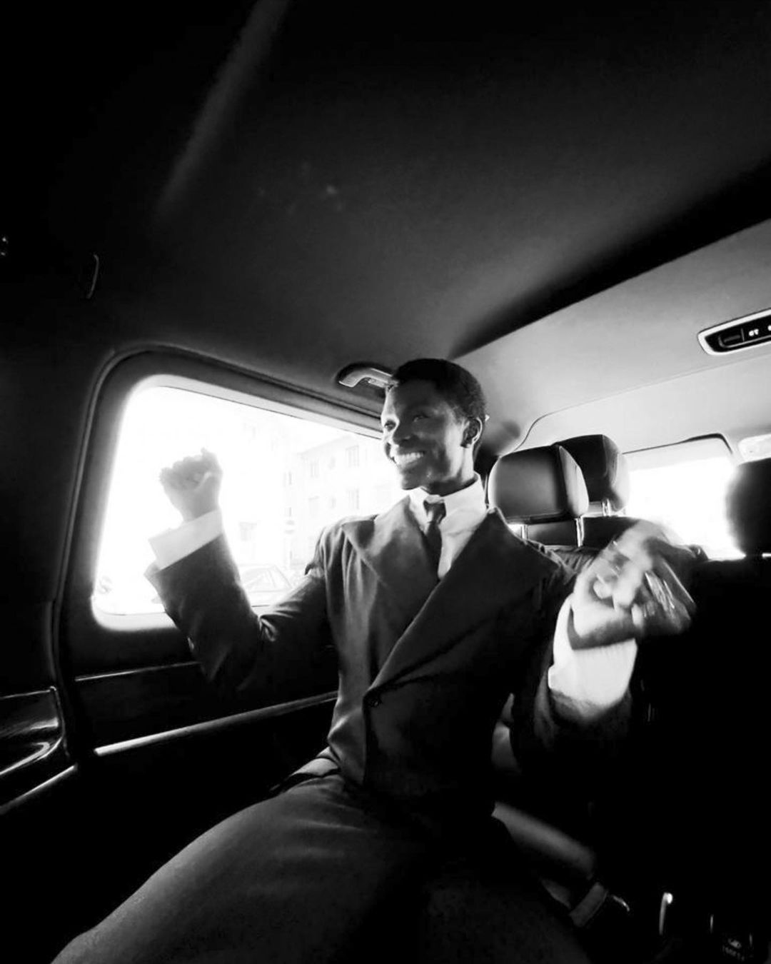 Happy African American man in suit riding in car celebrates with fists pumping, captured in monochrome tones with natural light.