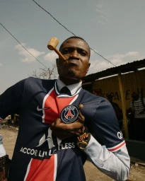 Man wearing a Paris Saint-Germain football jersey holding a corn cob pipe in his mouth while pointing at the club crest