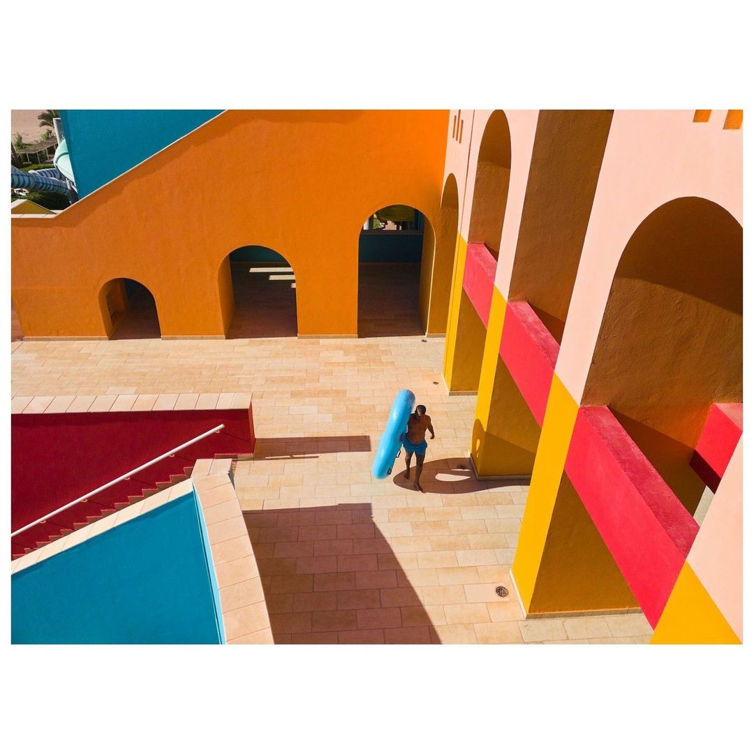 High-angle view of a man in blue swim trunks carrying an inflatable tube near a swimming pool and colorful modern architecture.