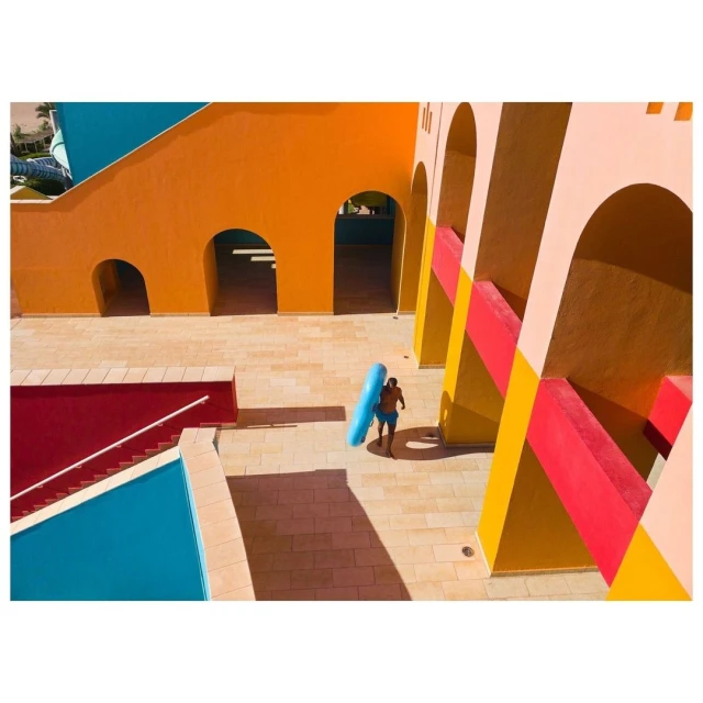 High-angle view of a man in blue swim trunks carrying an inflatable tube near a swimming pool and colorful modern architecture.