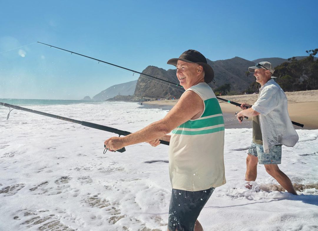 Two elderly men fishing in ocean surf on a sunny day; water, sand, and mountain backdrop fill the scene with recreational activity.