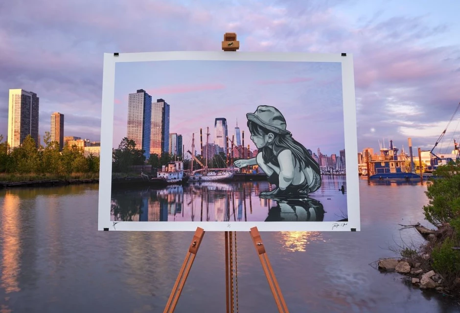 An outdoor photo showcases a print on an easel featuring a cityscape and a young girl, set against a backdrop of water and buildings.