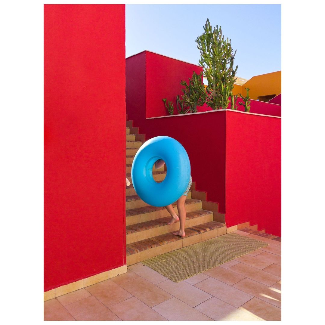 A child carries a blue inflatable ring up steps in front of a red building on a sunny day, presenting a vibrant and playful scene.