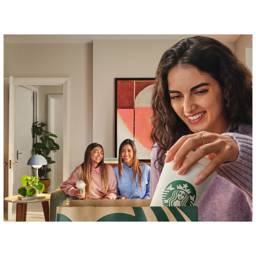 Three young women in a living room with Starbucks coffee in a takeout bag and beverage, focus on woman reaching into bag.