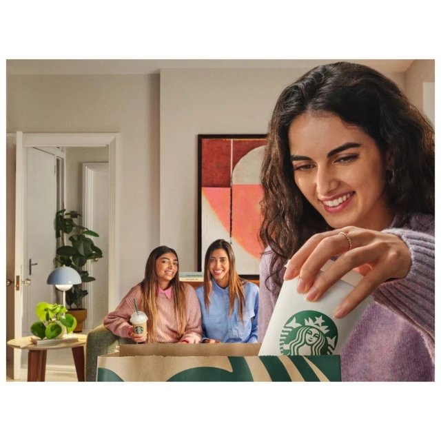 Three young women in a living room with Starbucks coffee in a takeout bag and beverage, focus on woman reaching into bag.