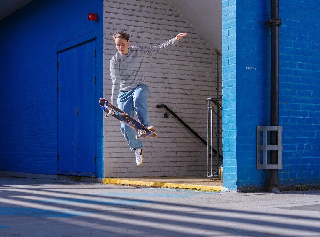 Female skateboarder in mid-air performing a trick with blue brick wall, doorway and concrete ground as the backdrop, outdoor setting.