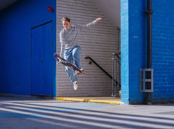 Female skateboarder in mid-air performing a trick with blue brick wall, doorway and concrete ground as the backdrop, outdoor setting.