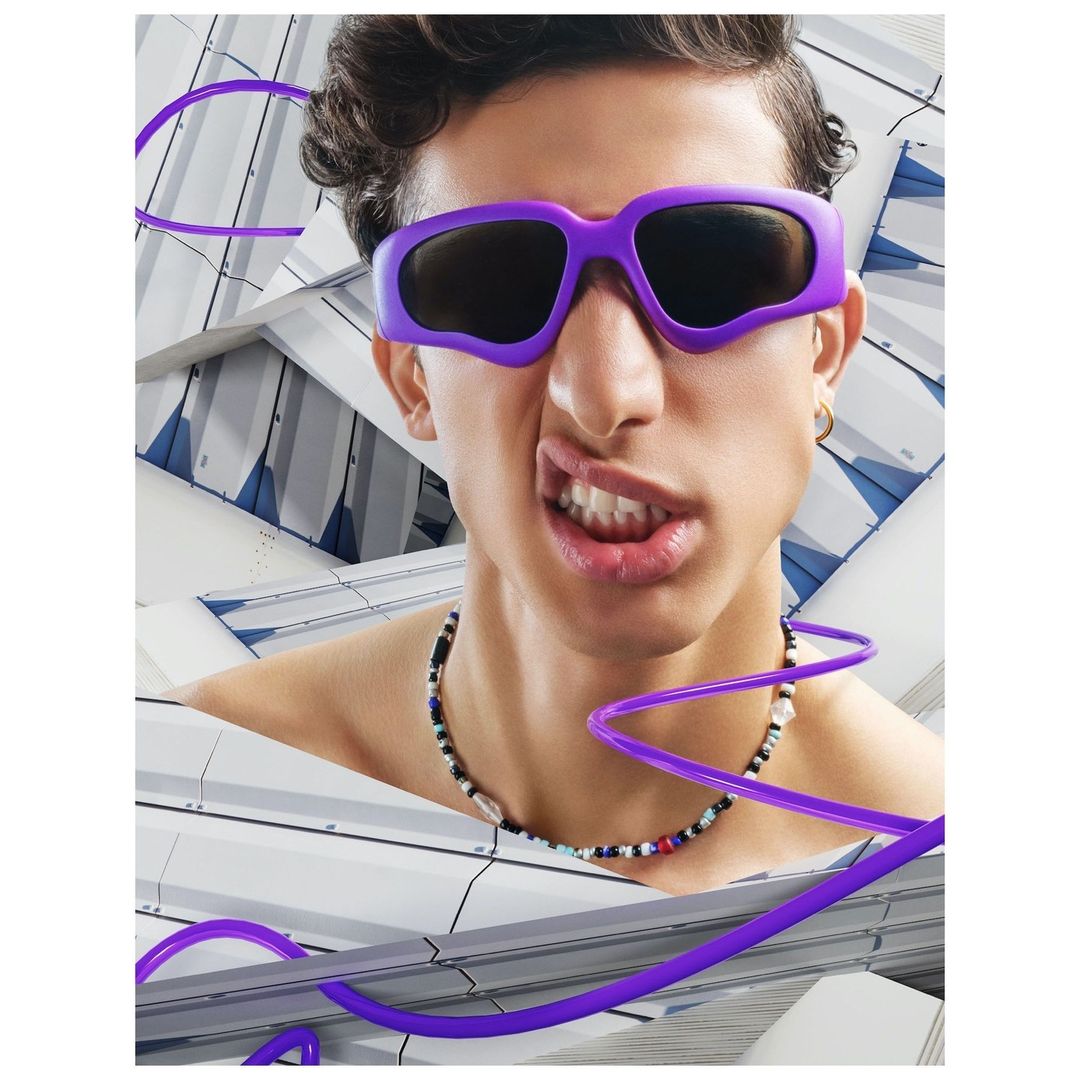 Close-up studio portrait of a young adult male with dark curly hair wearing purple sunglasses and necklace, set against a geometric background.