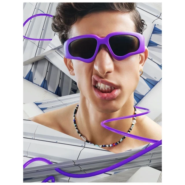 Close-up studio portrait of a young adult male with dark curly hair wearing purple sunglasses and necklace, set against a geometric background.