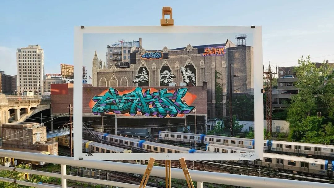 Graffiti art print on easel outdoors, featuring urban building and rail yard with trains, under clear sky and sunny lighting.