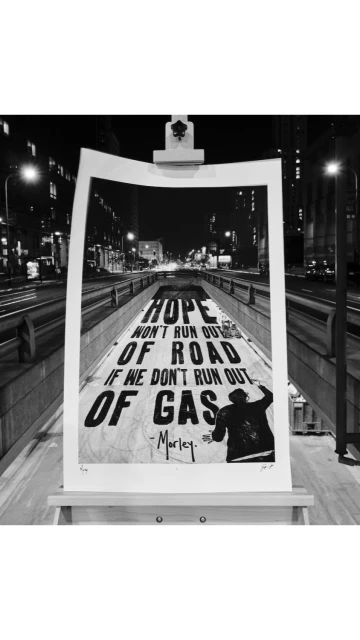 Black and white photograph of a poster on an easel displaying the text: “Hope won't run out of road if we don't run out of gas - Marley.