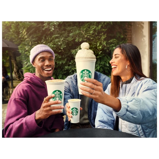 Two smiling adults hold Starbucks coffee cups outside, with a third cup with a knitted topper visible behind them in daylight.
