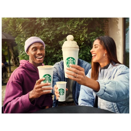 Two smiling adults hold Starbucks coffee cups outside, with a third cup with a knitted topper visible behind them in daylight.