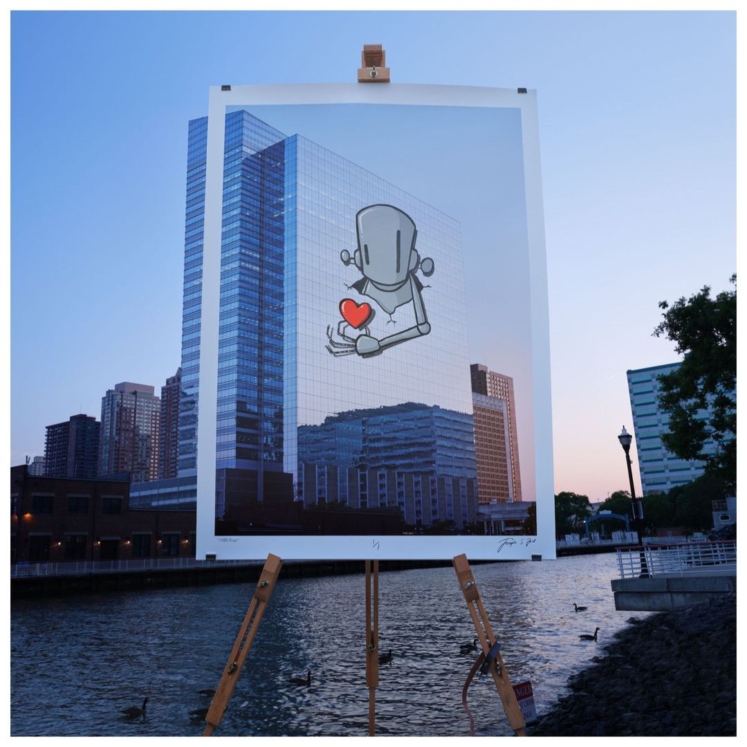 Cartoon robot art print on an easel by a river, set against a backdrop of modern buildings at dusk with a blue sky overhead.
