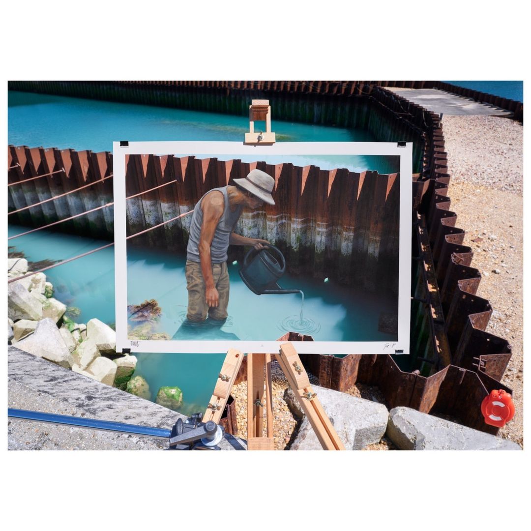 Painting of a man watering a body of water near a retaining wall is presented on an easel outdoors near rocks and turquoise water.