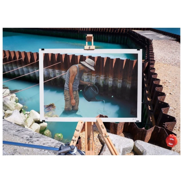 Painting of a man watering a body of water near a retaining wall is presented on an easel outdoors near rocks and turquoise water.