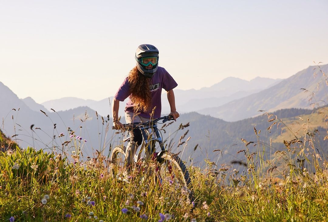 Female mountain biker rides downhill on a mountain bike through a grassy, wildflower-covered field with a mountain range backdrop.