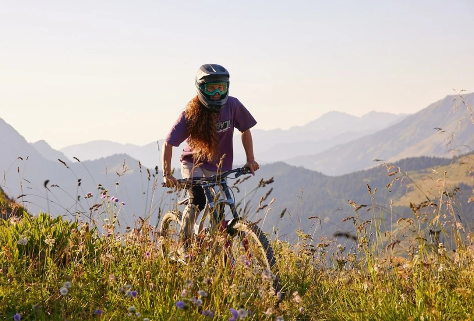 Female mountain biker rides downhill on a mountain bike through a grassy, wildflower-covered field with a mountain range backdrop.