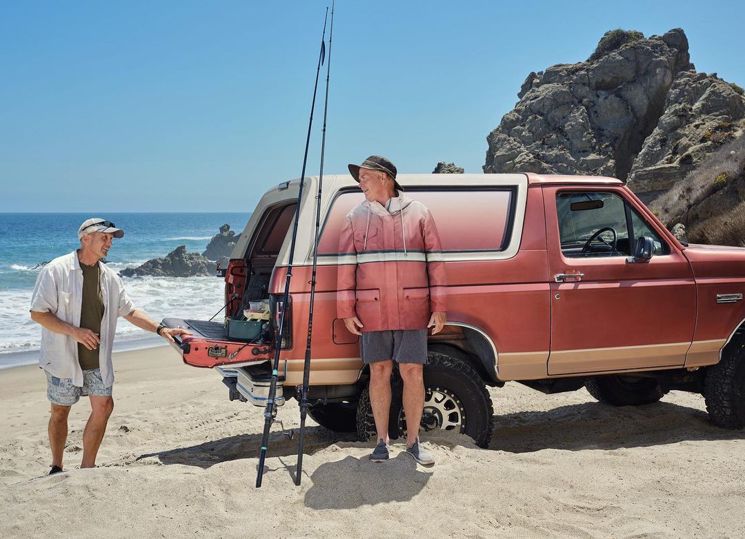 Two men near a red SUV on a sandy beach prepare for fishing with fishing rods. Ocean and rock formations in background on sunny day.