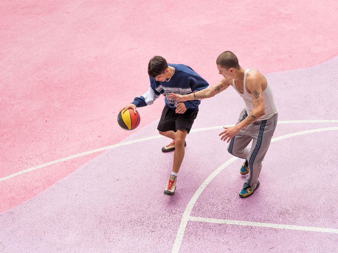 Two young men playing basketball on an outdoor pink court. One dribbles while the other defends in casual athletic wear and natural lighting.