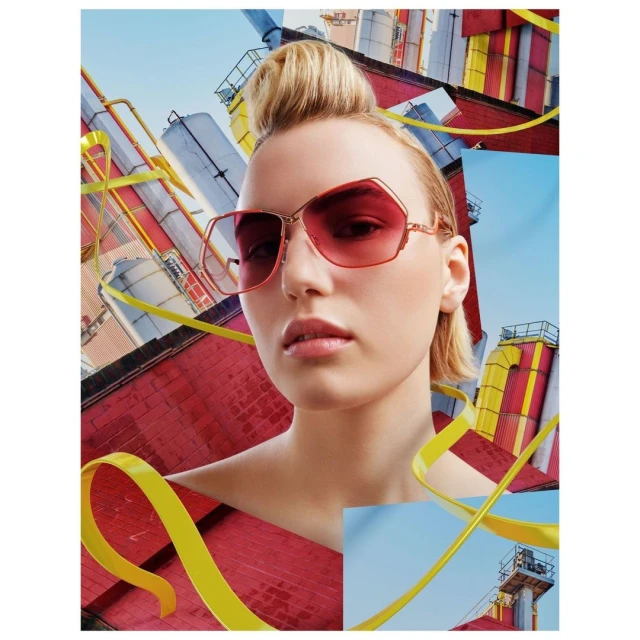A woman with pink sunglasses poses against a colorful industrial backdrop with yellow ribbons for a fashion or beauty advertisement.