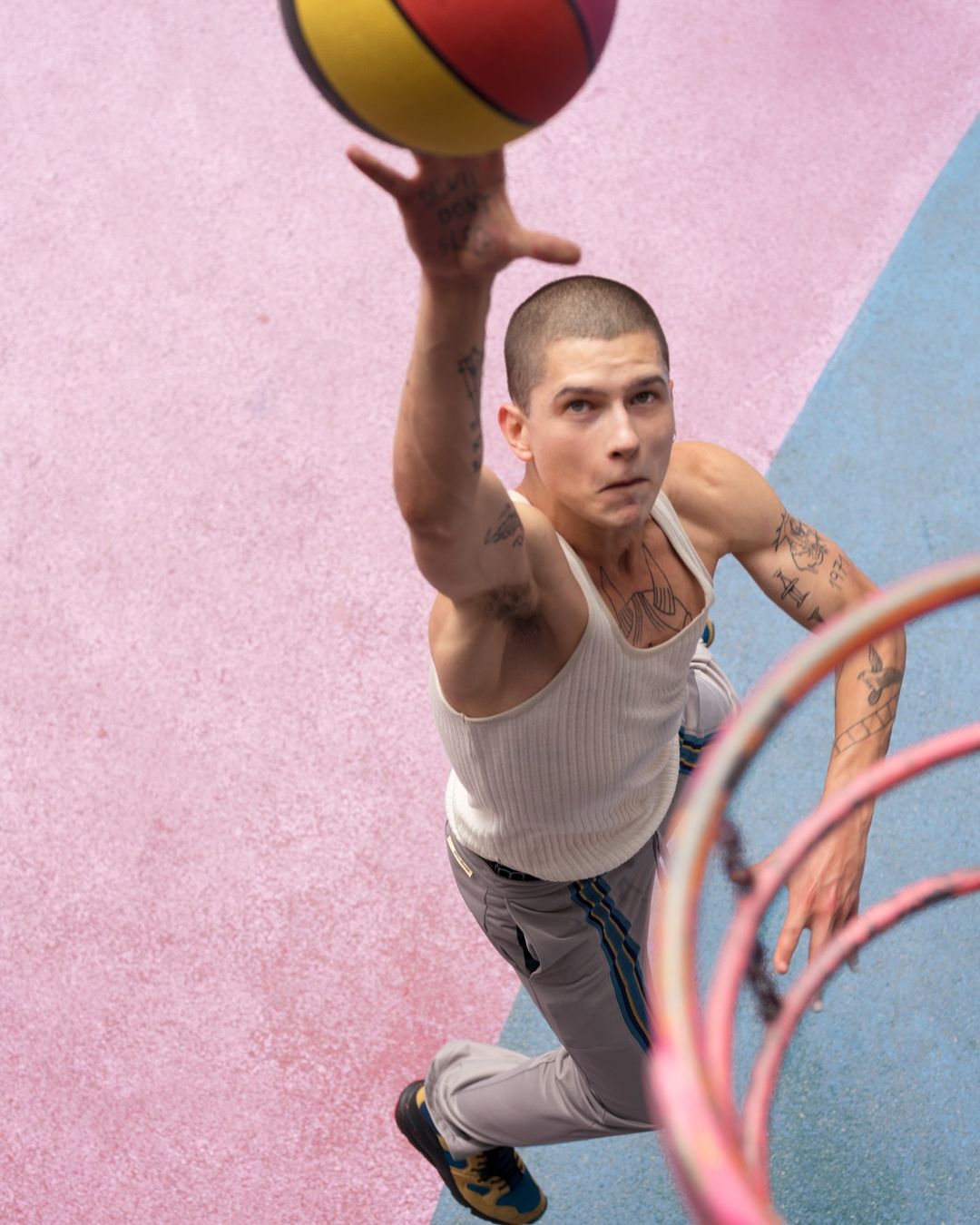 Low-angle shot of a young adult male with short hair, wearing a tank top, reaching up to catch a basketball on a pink and blue court.