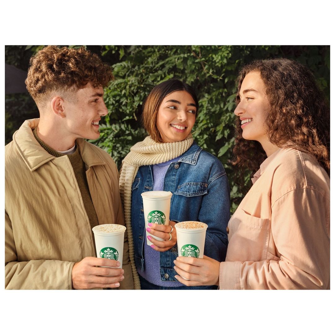 Three friends, two women and a man, smiling while holding Starbucks coffee cups outdoors in natural light.