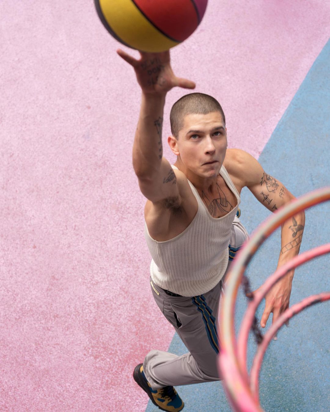 Athletic man in tank top throwing basketball toward hoop on colorful outdoor court with pink and blue sections in worm's-eye view.