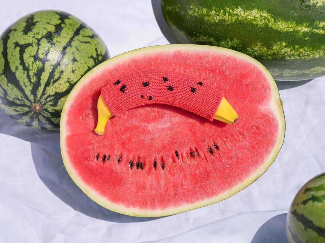 A sliced watermelon with a banana in a red knitted cover, creating a humorous visual pun, set against a white fabric backdrop.