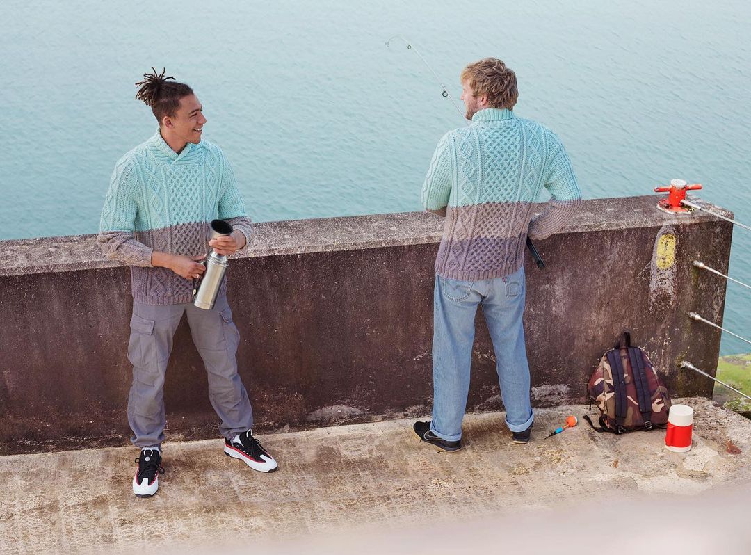 Two men in matching sweaters stand on a concrete structure overlooking a body of water on a cloudy day, captured in an eye-level shot.
