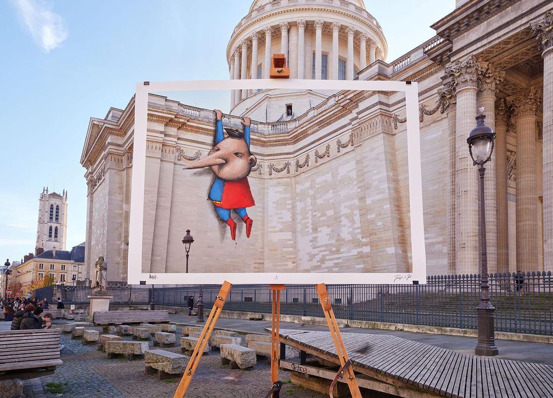 Outdoor street art display featuring a cartoon superhero graphic propped in front of a classical building, with a gathering of people.