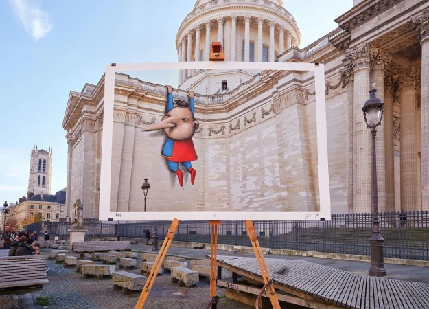 Outdoor street art display featuring a cartoon superhero graphic propped in front of a classical building, with a gathering of people.