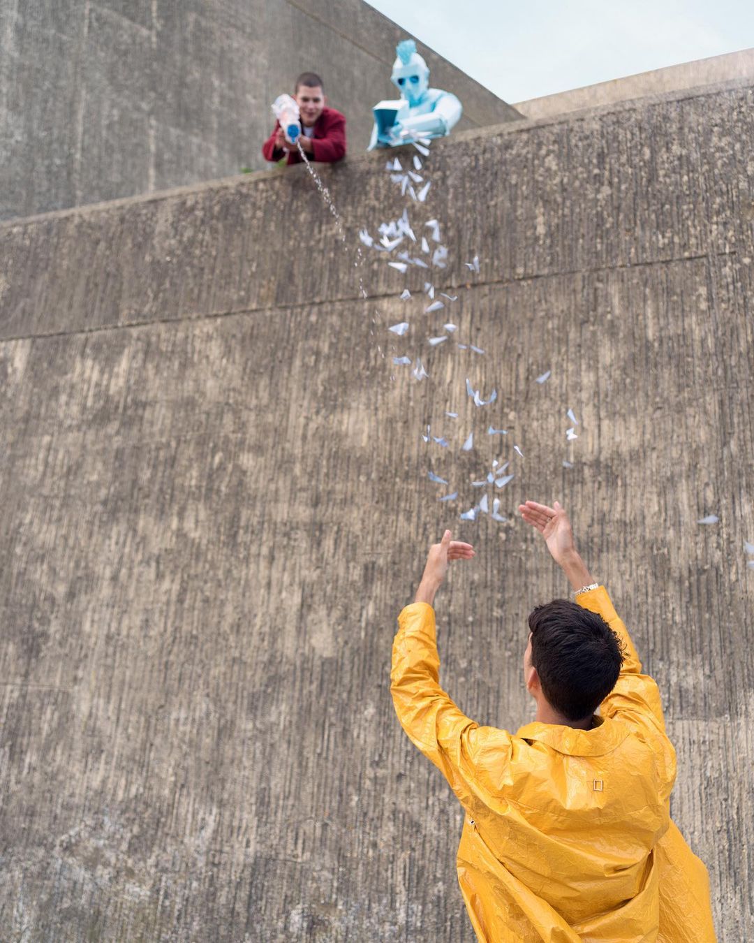 Three people near a concrete wall, two are on top shooting paper confetti down to the third person wearing a yellow raincoat.