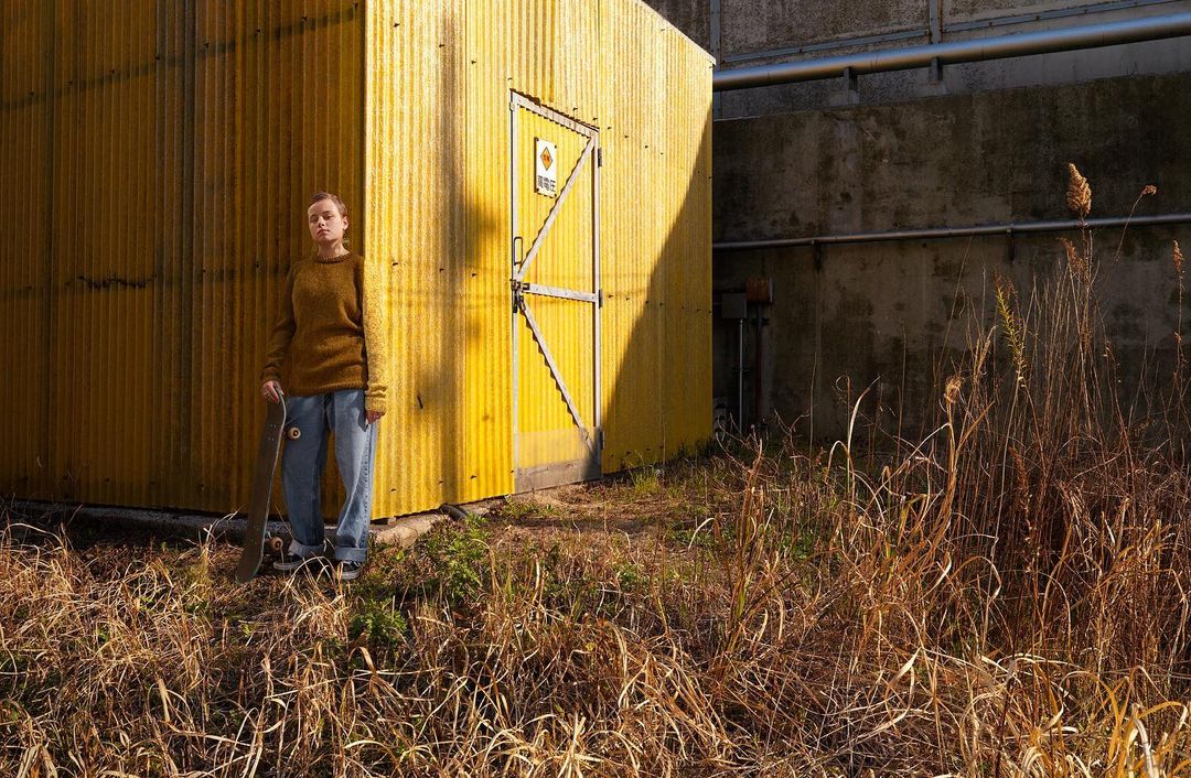 Young woman standing near a yellow building with a skateboard in hand on a sunny day in a grassy, urban outdoor environment.