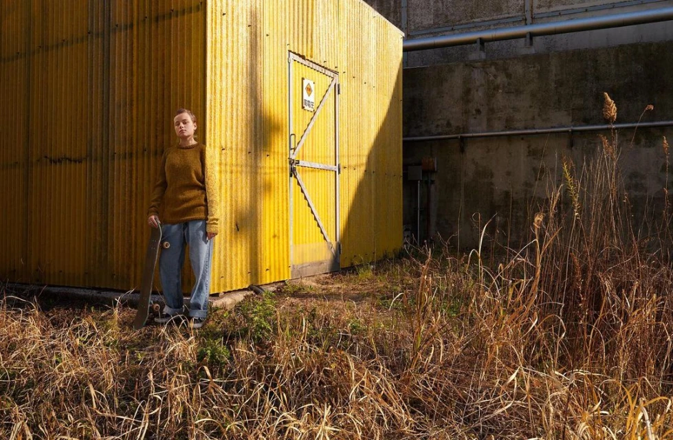 Young woman standing near a yellow building with a skateboard in hand on a sunny day in a grassy, urban outdoor environment.