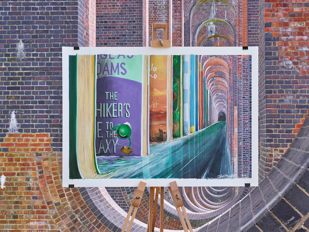 Painting on an easel depicts a row of colorful books, one featuring 'The Hitchhiker's Guide to the Galaxy,' set against a brick viaduct.