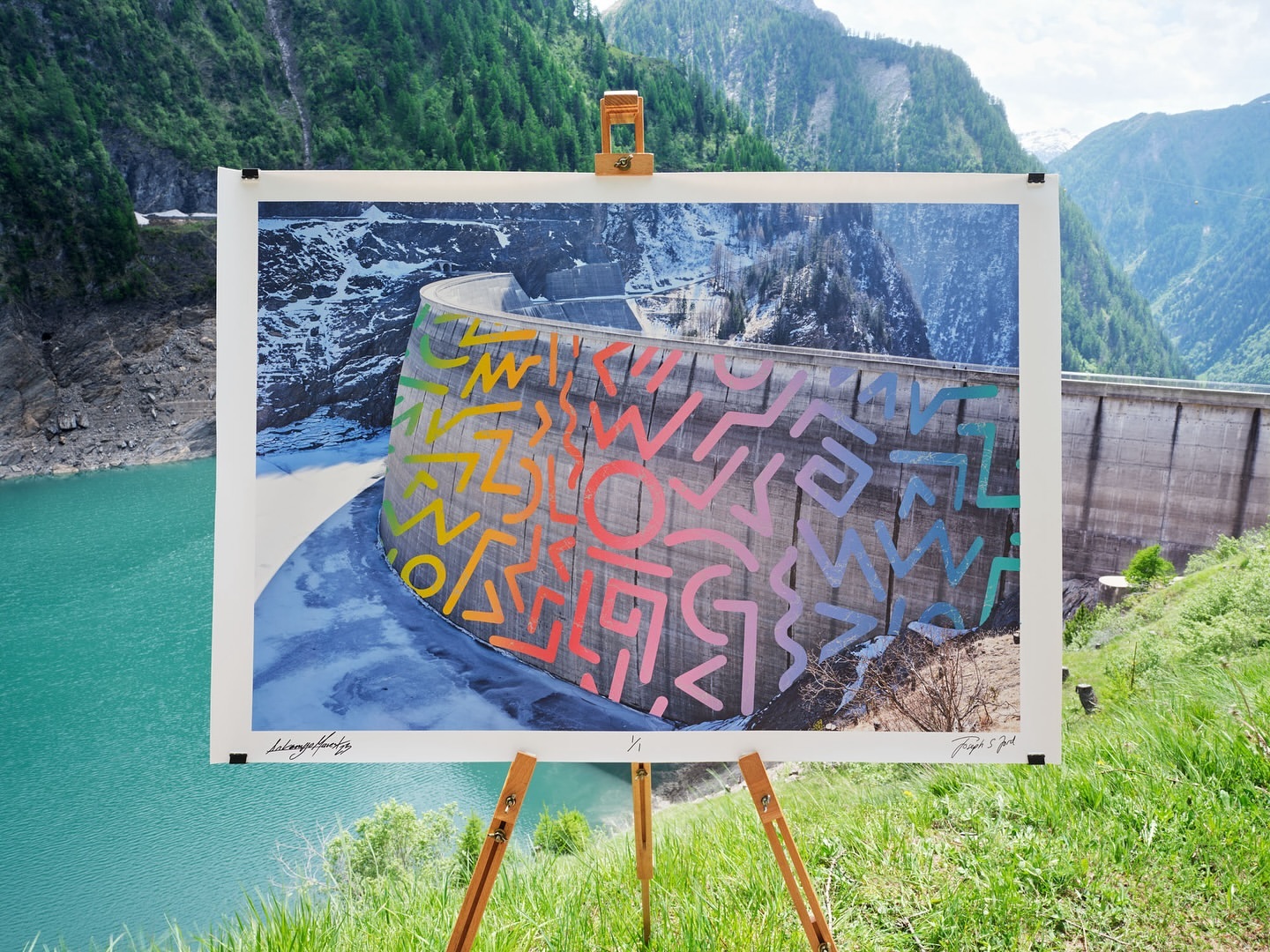 Artwork featuring a concrete dam with colorful, geometric designs, displayed on an easel against a mountain and lake backdrop.