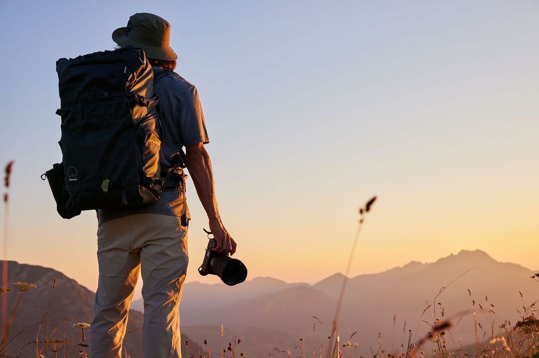 Person with backpack and camera looking at mountain range during sunset. Outdoor photography in nature, travel and adventure concept.