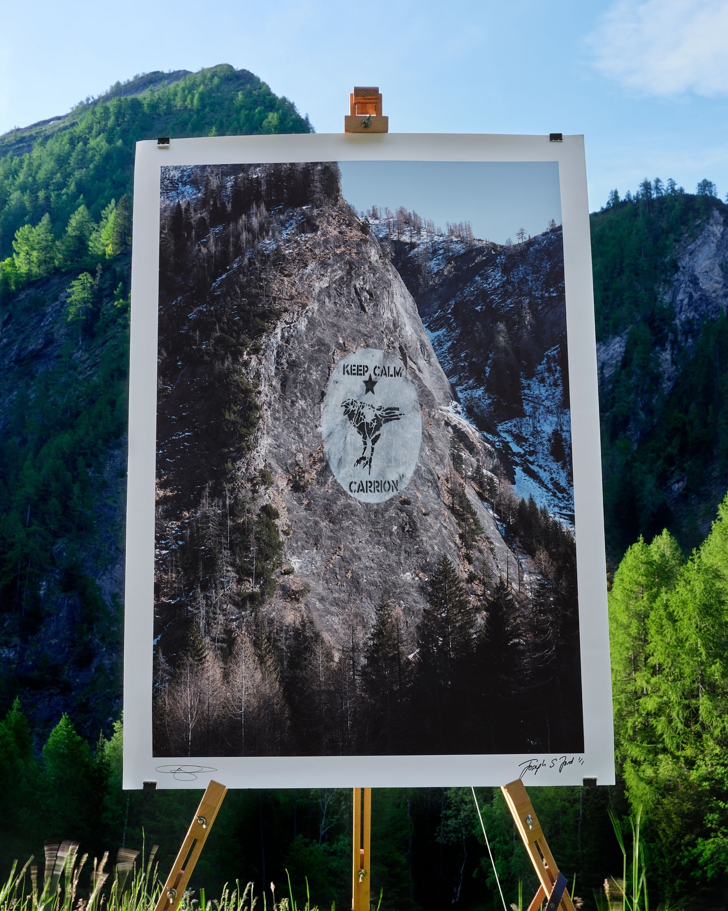 Art print of mountain landscape on a wooden easel in an outdoor setting with a forest and mountain range in the background.