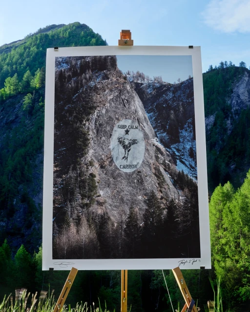 Art print of mountain landscape on a wooden easel in an outdoor setting with a forest and mountain range in the background.