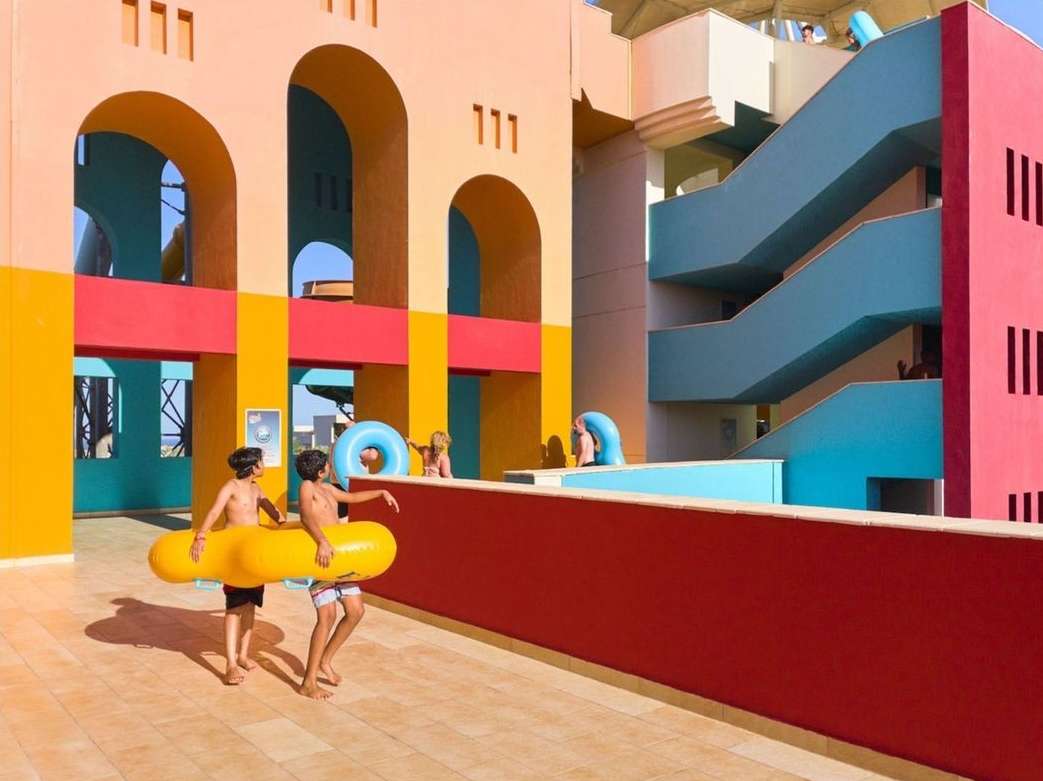 Two children carry a large yellow inflatable pool toy at a resort with colorful architecture and stairs on a sunny day outdoors.