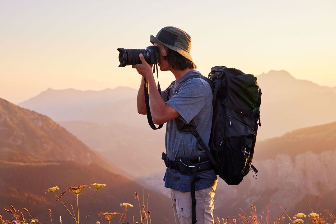 A photographer in a hat with a large camera takes a photo on a mountain ridge at sunset, wearing a grey shirt and a large backpack.