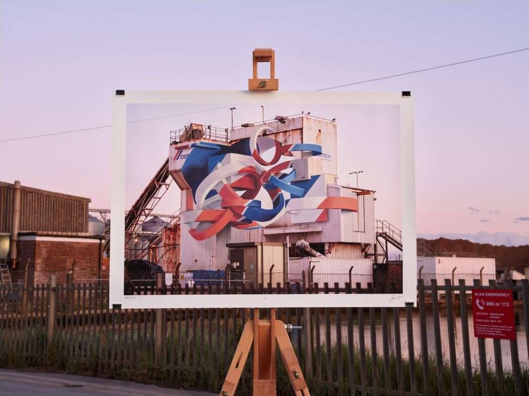 Wooden easel presents a large photo print of a white industrial building with abstract graffiti against a pink and purple sky at dusk.