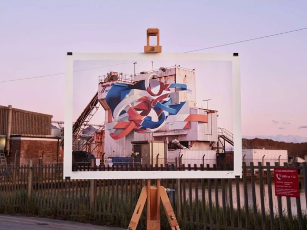 Wooden easel presents a large photo print of a white industrial building with abstract graffiti against a pink and purple sky at dusk.